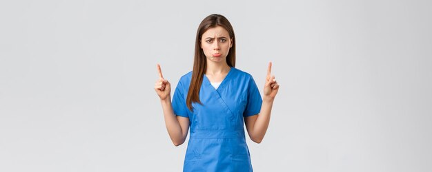 Healthcare workers prevent virus insurance and medicine concept Upset sobbing cute nurse or doctor in blue scrubs pouting feel uneasy pointing fingers up at unpleasant bad news show banner