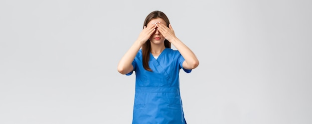 Healthcare workers prevent virus insurance and medicine concept Young woman in blue scrubs doctor or nurse close eyes with hands blindfolded medicine standing grey background