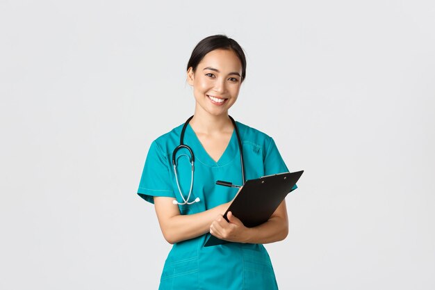 ウイルス検疫キャンペーンのコンセプトを防ぐ医療従事者毎日の健康診断中にクリップボードを持って陽気なフレンドリーなアジアの女性医師医師立っている白い背景