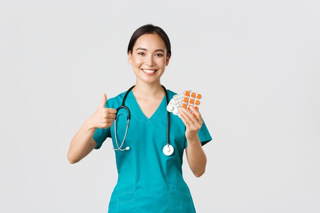 Медицинские работники предотвращают концепцию вирусной карантинной кампании Уверенно улыбающаяся азиатская медсестра-врач в скрабах рекомендует лекарства показать большой палец вверх и таблетки на белом фоне