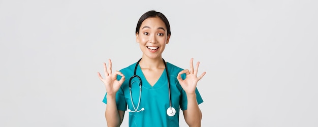 Healthcare workers, preventing virus, quarantine campaign concept. Excited and pleased asian female doctor, nurse in scrubs with stethoscope, showing okay gesture, white background.