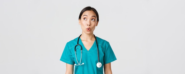 Healthcare workers, preventing virus, quarantine campaign concept. Impressed and amazed asian female doctor, nurse in medical scrubs looking upper left corner pleased, white background