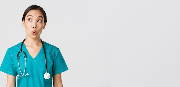 Healthcare workers, preventing virus, quarantine campaign concept. Impressed and amazed asian female doctor, nurse in medical scrubs looking upper left corner pleased, white background.