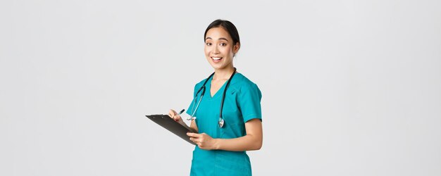 의료 종사자, 바이러스 예방, 검역 캠페인 개념. 웃고 있는 귀여운 아시아 여성 간호사, 수술실 인턴, 클립보드에 글을 쓰고, 종이를 채우고, 카메라가 만족스러운 흰색 배경을 보고 있습니다.