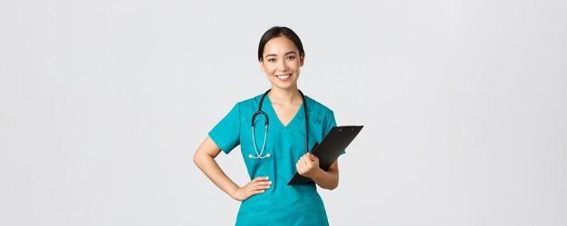 医療従事者、ウイルスの予防、検疫キャンペーンのコンセプト。笑顔の楽しいアジアの女性医師、スクラブを着用し、クリップボード、白い背景を保持している検査中の医師。