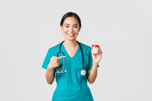 바이러스 검역 캠페인 개념을 예방하는 의료 종사자들은 수술복을 입은 미소 짓는 예쁜 아시아 여성 의사 간호사가 비타민을 보여주고 약을 가리키며 흰색 배경의 약을 추천합니다