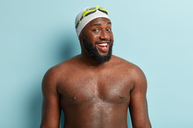 Healthy black man with athletic body shape, being professional swimmer, comes out of water, feels relaxed and full of energy, wears swimming cap, goggles, isolated on blue wall