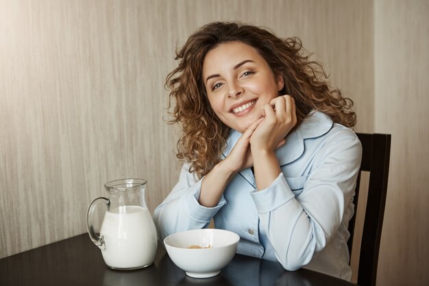 家族の輪で健康的な朝食。牛乳と一緒に穀物を食べながら手に寄りかかって巻き毛の美しい若い母親、幸せな笑顔、夫とのチャット