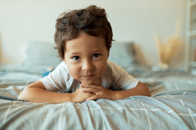 Healthy charming 3 year old Latin toddler lying on rumpled sheets with hands clasped in front of him, having curious play fun facial expression