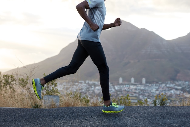 Free photo healthy dark skinned man in action, runs along road near mountains, wears comfortable sneakers, casual clothes, has sporty body. fast male athlete poses against sky. racing competition