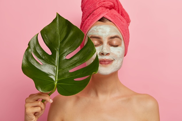 Healthy European woman applies facial mask for rejuvenation and removing pores, holds green leave, stands with closes eyes, naked body, wrapped towel on head, models indoor. Cosmetology, beauty