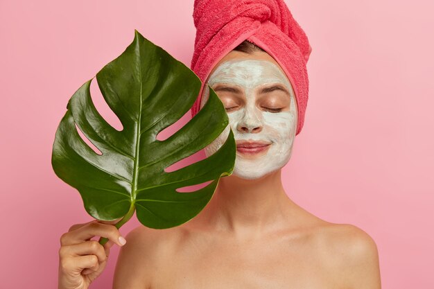 Healthy European woman applies facial mask for rejuvenation and removing pores, holds green leave, stands with closes eyes, naked body, wrapped towel on head, models indoor. Cosmetology, beauty