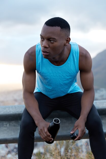Healthy fatigue dark skinned man takes break after outdoor workout, holds bottle of energy drink, has pensive look aside, models outside alone, refreshes with water, tries to rejuvenate, being strong