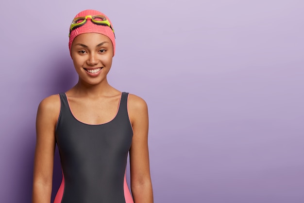 Healthy female swimmer wears pink cap, goggles, swimming costume, prepares for workout in pool