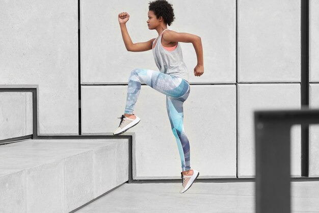 Healthy fit woman runs up on stairs, wears comfortable clothes and trainers, has jogging workout, does sport in urban environment, being fast, poses sideways. Wellness and determination concept