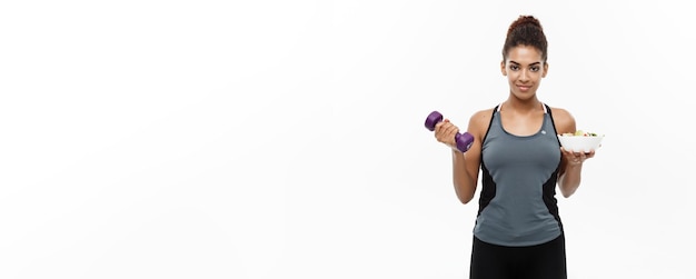 Healthy and Fitness concept Beautiful sporty African American on diet holding dumbbell and fresh salad on hands Isolated on white studio background