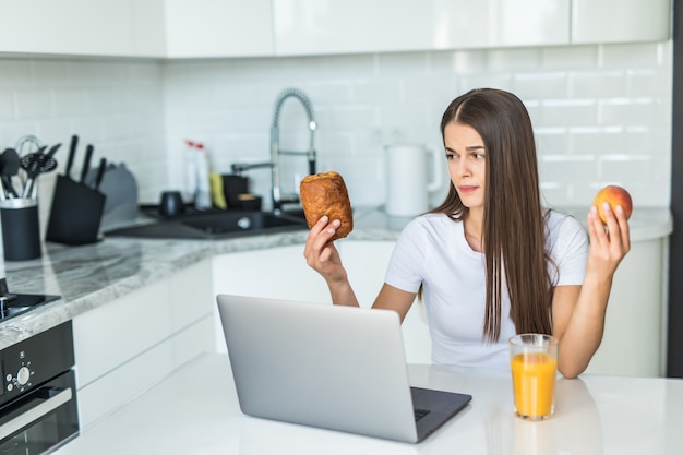 Healthy food concept. Hard choice. Yound sporty woman is choosing between healthy food and sweets while standing on light kitchen.