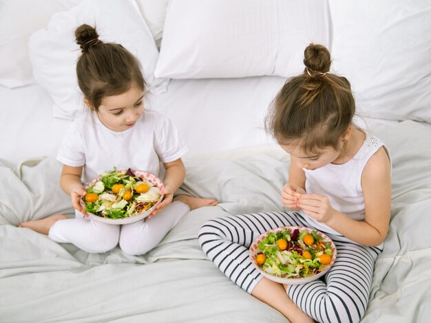 自宅で健康食品。ベッドの上の寝室で果物と野菜を食べて幸せな2人のかわいい子供。子供とティーンエイジャーのための健康食品。