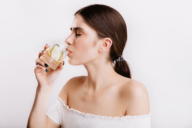 Healthy girl with clear skin drinks water with lemon and cucumber in morning. Shot of beautiful model without makeup on white wall.