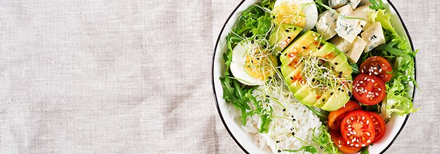 Healthy green vegetarian buddha bowl lunch with eggs, rice, tomato, avocado and blue cheese on table.