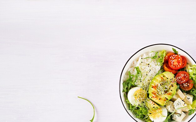 Healthy green vegetarian buddha bowl lunch with eggs, rice, tomato, avocado and blue cheese on table.