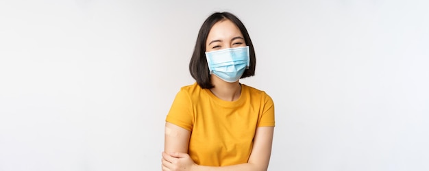 医療用フェイスマスクの健康で幸せなアジアの女性は、covid19ワクチン接種がオミクロンデルタバリアントの背景からワクチン接種された後、肩にバンドエイドを持っています