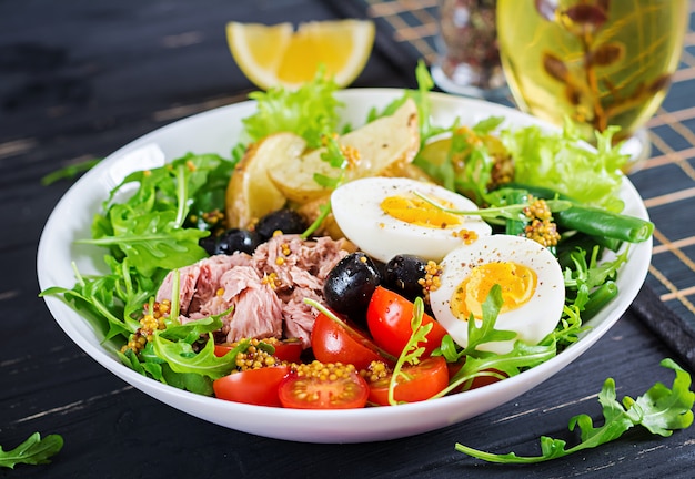 Healthy hearty salad of tuna, green beans, tomatoes, eggs, potatoes, black olives close-up in a bowl on the table.
