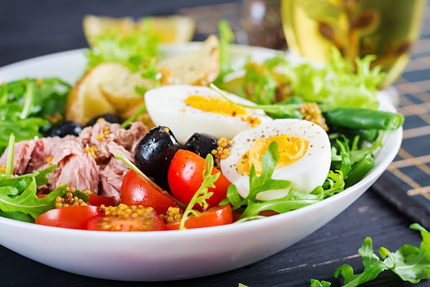 Healthy hearty salad of tuna, green beans, tomatoes, eggs, potatoes, black olives close-up in a bowl on the table