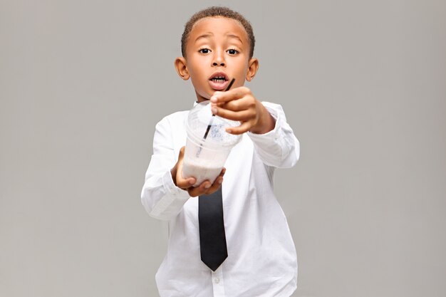 Healthy lifestyle, food, beverage and childhood concept. Picture of emotional excited African American schoolboy in uniform holding plastic jar with fruit milkshake, offering you to have some