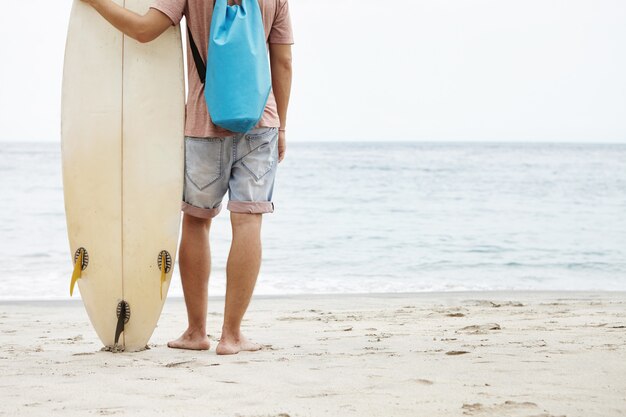 Healthy lifestyle and leisure concept. Rear cropped view of Caucasian tourist standing barefooted on sandy beach and holding white surfboard, facing calm and peaceful ocean