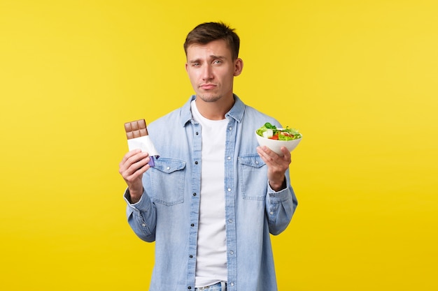 Концепция здорового образа жизни и эмоций людей. Мрачный красивый кавказский парень показывает салат в миске и шоколадном батончике, морщась от того, что неохотно ест диетическую пищу, стоя на желтом фоне