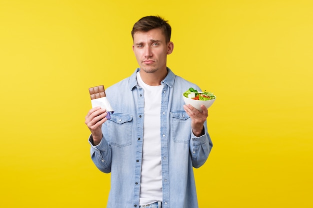 Healthy lifestyle and people emotions concept. Gloomy handsome caucasian guy showing salad in bowl and candy bar, grimacing as reluctant eat diet food, standing yellow background