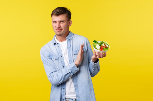 Healthy lifestyle, people and food concept. Disgusted and disappointed blond man grimacing from aversion and showing refusal to bowl with salad, dislike staying on diet, yellow background.