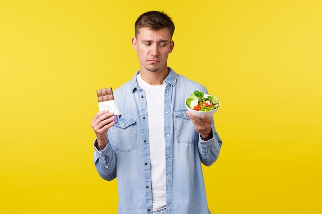 Healthy lifestyle, people and food concept. Displeased and reluctant handsome sad guy want eat candy bar and looking with disgust at bowl with salad, sit on diet, standing yellow background.