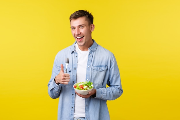 Healthy lifestyle, people and food concept. Happy smiling man showing thumbs-up satisfied with delicious breakfast, eating salad, being on diet, trying stay fit, standing yellow background.