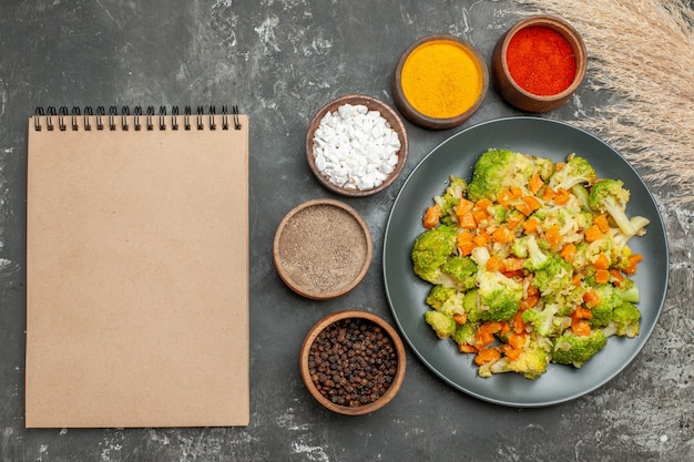 Healthy meal with brocoli and carrots on a black plate and spices next to notebook on gray table