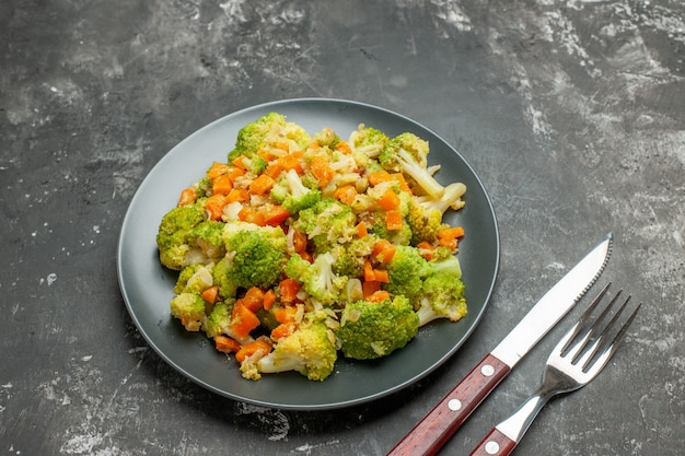 Pasto sano con broccoli e carote su un piatto nero con forchetta e coltello sul tavolo grigio