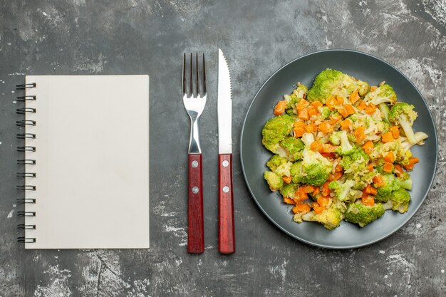 Foto gratuita pasto sano con broccoli e carote su un piatto nero con forchetta e coltello accanto al taccuino