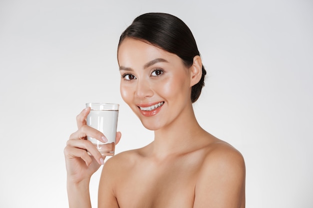 Healthy portrait of young happy woman with hair in bun drinking still water from transparent glass, isolated over white