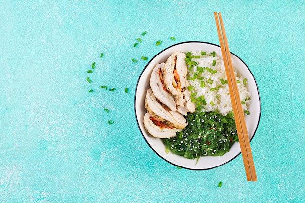 Healthy salad in a white bowl, chopsticks. Chicken rolls, rice, chuka and green onion. Blue table. Asian cuisine. Top view