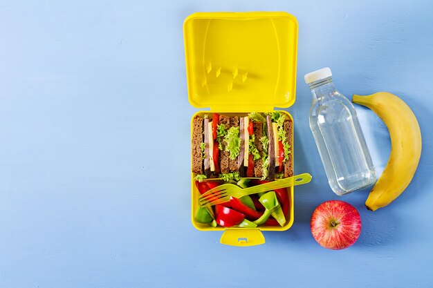 Здоровая коробка школьного обеда с сандвичем говядины и свежими овощами, бутылкой воды и плодоовощами на голубой таблице. Вид сверху. Плоская планировка