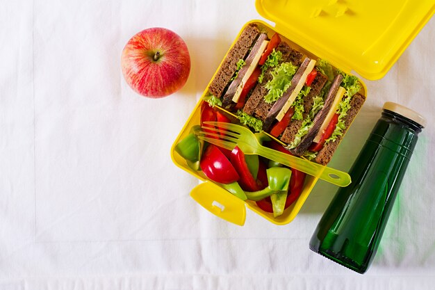 Здоровая коробка школьного обеда с сандвичем говядины и свежими овощами, бутылкой воды и плодоовощами на белой таблице. Вид сверху. Плоская планировка