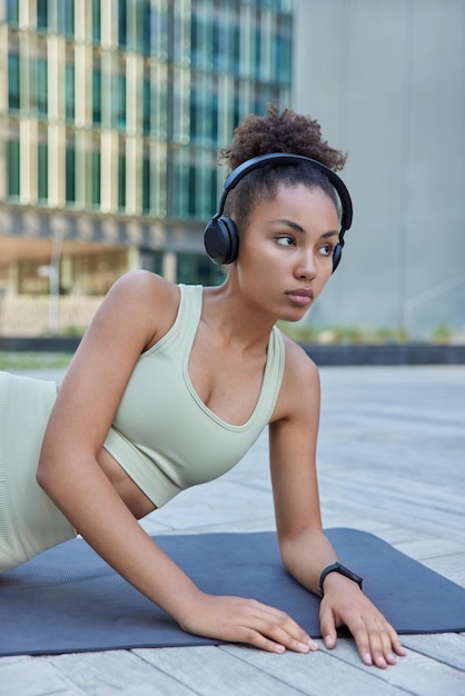 Healthy slim woman with curly hair dressed in sportswear lies on fitness mat rests after cardio training listens music via wireless headphones