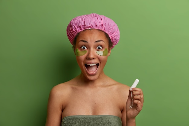Modello femminile sorridente sano avvolto in un asciugamano da bagno, indossa cerotti di bellezza verdi sotto gli occhi
