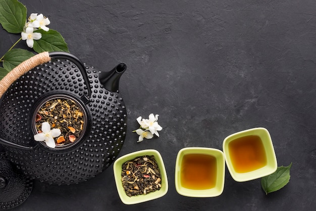 Healthy tea with aromatic dry tea in bowls and teapot on black surface