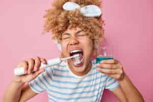 Free photo healthy teeth and care concept cheerful curly haired woman brushes teeth with electric toothbrush holds glass of blue mouthwash takes care of oral cavity wears headband and striped t shirt