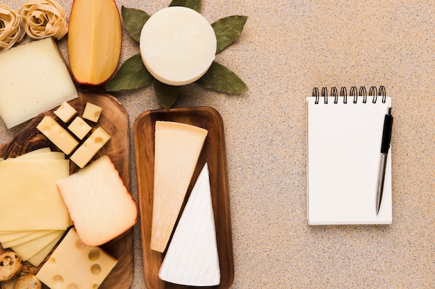 Healthy types of cheeses on wooden plate with blank white notepad and pen