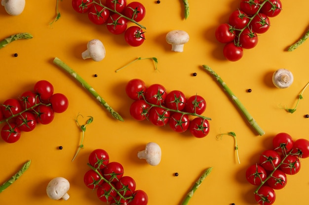 Healthy vegetables used for cooking purposes. Ripe red cherry tomatoes, sprouts of asparagus, white champignons and peppercorns isolated on yellow background. Products from grocery store or market