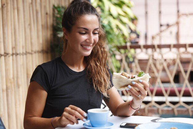 日焼けした健康な女性、カフェテラスでTシャツに座って、朝食を食べ、コーヒーを飲みます。