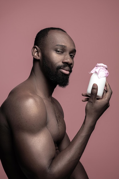 Sano giovane africano holging piccola bottiglia di latte su uno sfondo rosa studio. ritratto ravvicinato in stile minimalista di un giovane uomo afro felice nudo